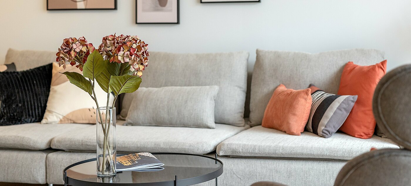 Se ve el respaldo de un sofá y una mesita de cristal con flores en un jarrón delante