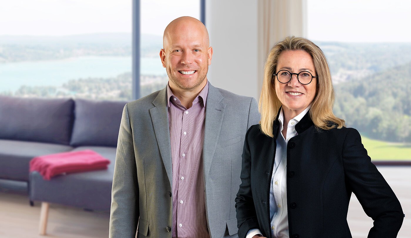 Das Mr. Lodge Team der Filiale in Rottach-Egern Petra Berger und Alexander Thamm, das Team der Mr. Lodge Filiale am Tegernsee, vor traumhafter Seelandschaft – Ihre Ansprechpartner für exklusive möblierte Immobilien in der Region.